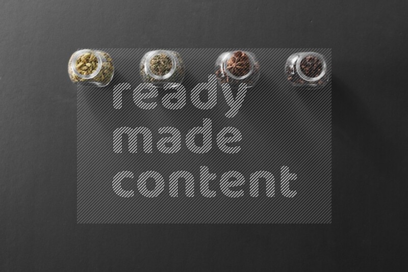 4 glass spice jars full of cloves, cardamom, basil and star anise on black flooring