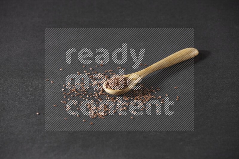 A wooden spoon full of flaxseeds and seeds spread beside it on a black flooring