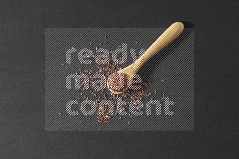 A wooden spoon full of flaxseeds and seeds spread beside it on a black flooring