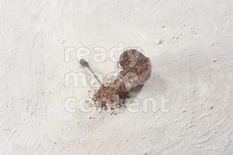 A glass spice jar full of flax seeds flipped with a metal spoon full of the seeds on a textured white flooring