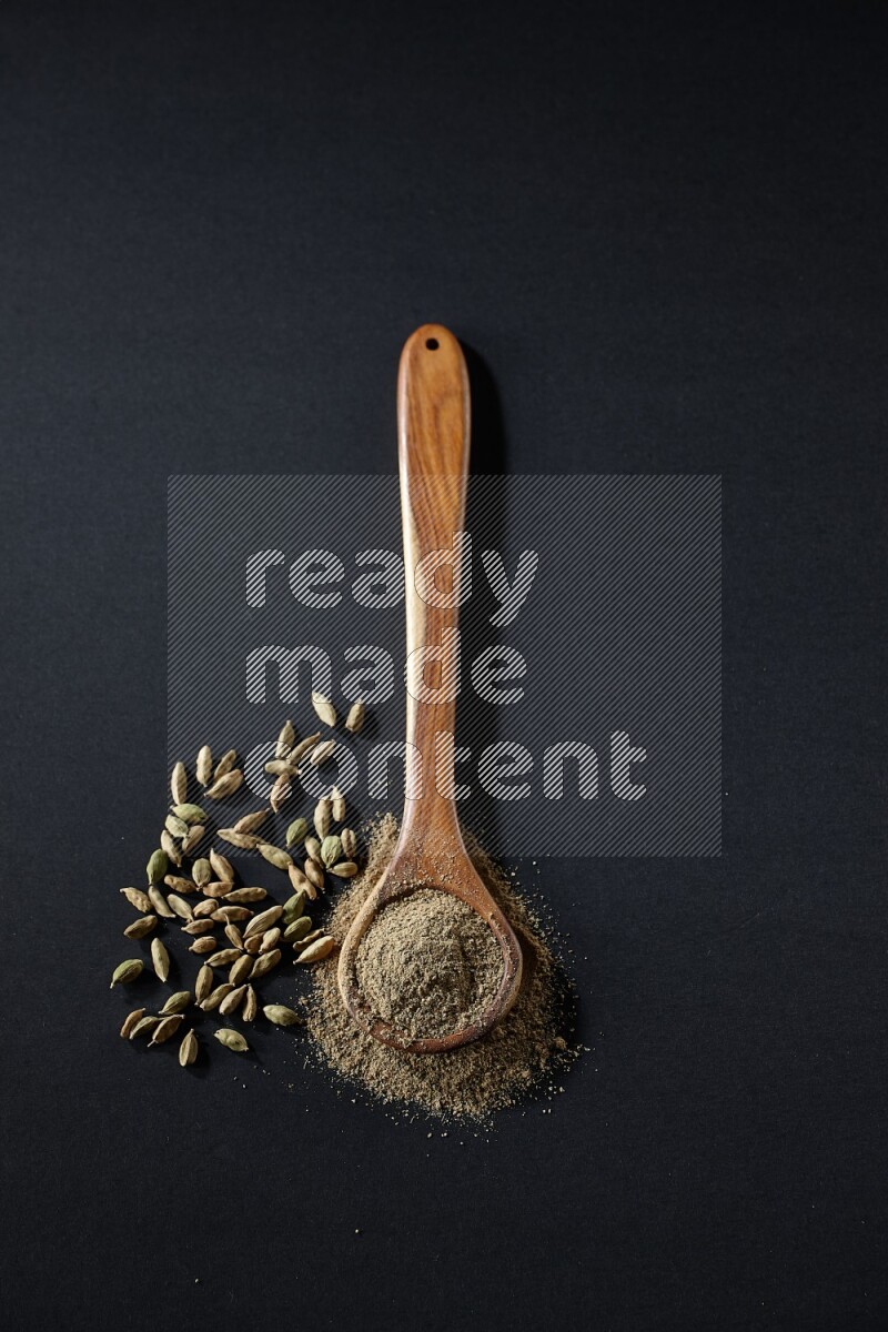 A wooden ladle full of cardamom powder and cardamom seeds beside it on black flooring