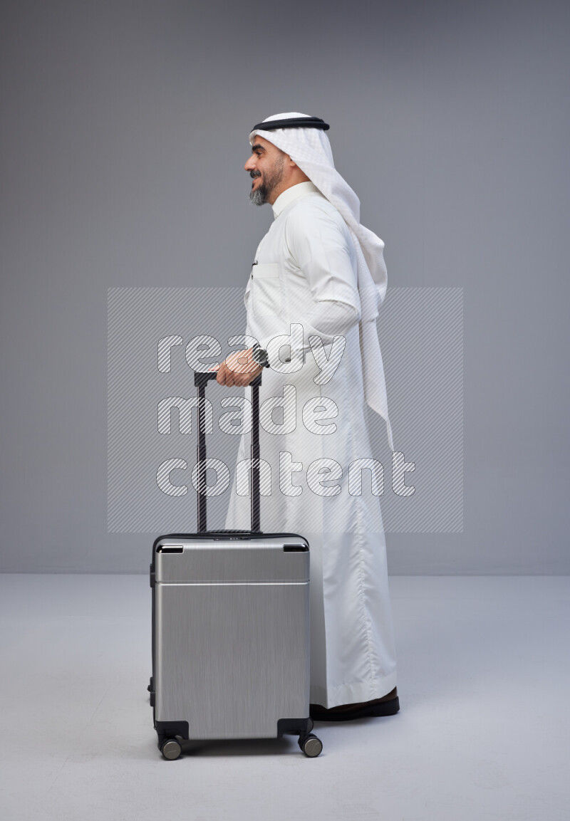 Saudi man wearing Thob and white Shomag standing holding Travel bag on Gray background