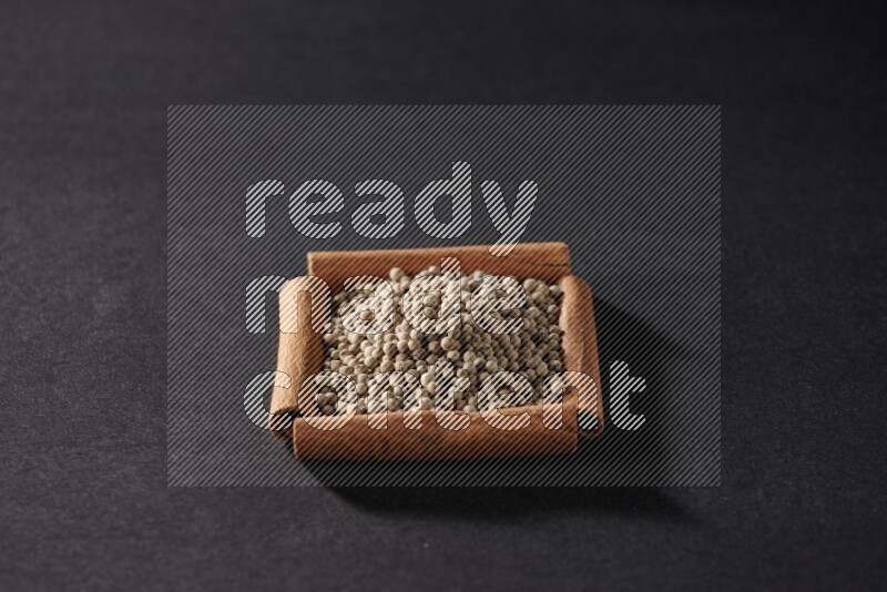 أربعة من أعواد القرفة مكونة شكل مربع بداخلها حبوب الفلفل الأبيض علي خلفية سوداء