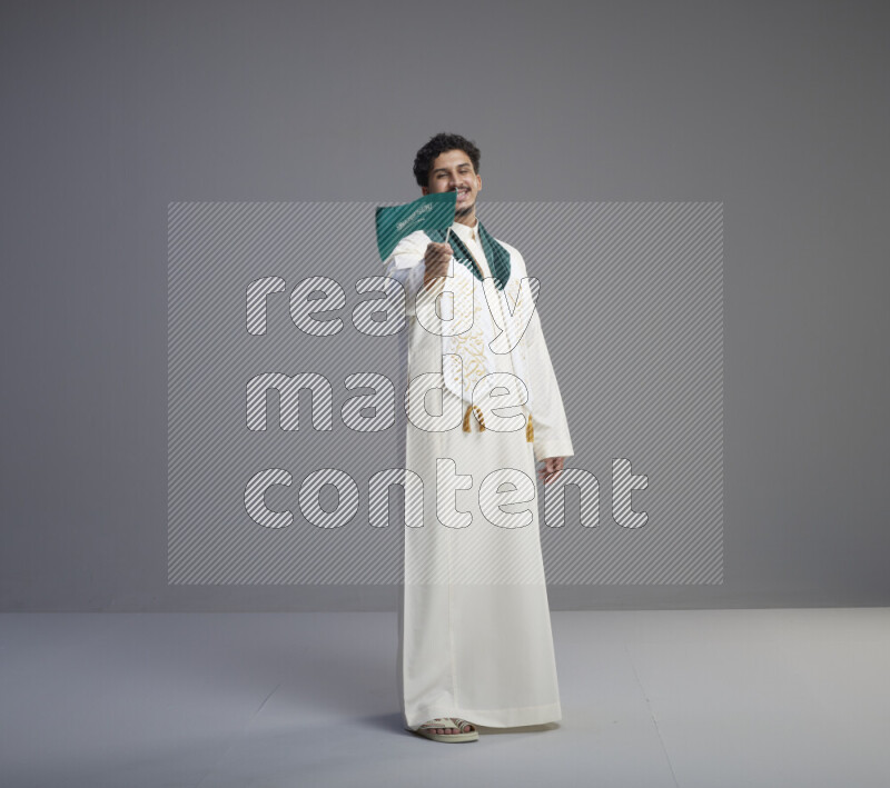A Saudi man standing wearing thob and saudi flag scarf and holding small saudi flag on gray background