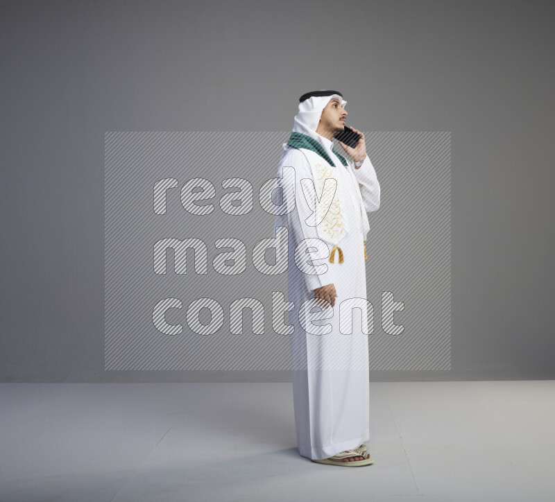 A saudi man standing wearing thob and white shomag with flag scarf on his neck talking on phone on gray background