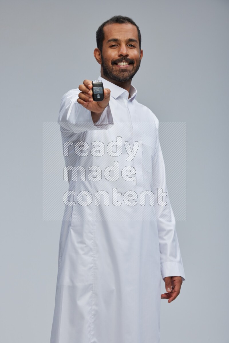 Saudi man wearing thob and shomag standing holding car key on gray background