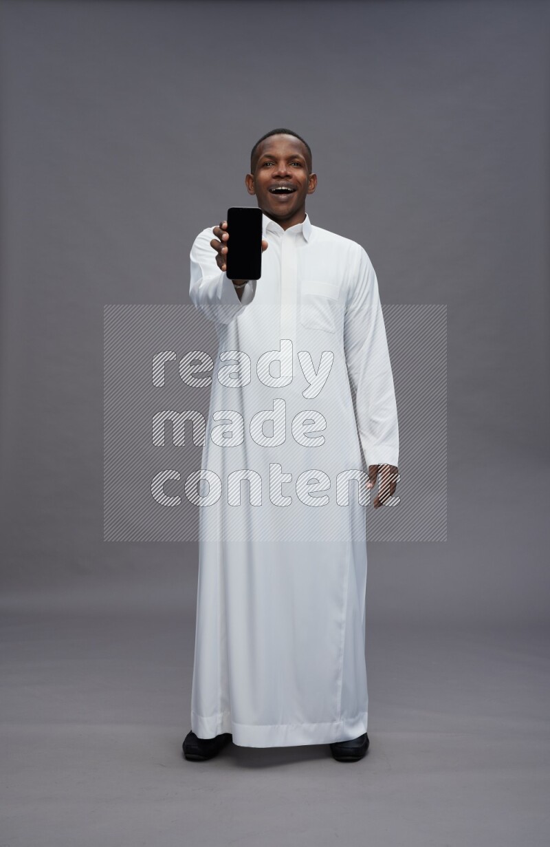 Saudi man wearing thob standing showing phone to camera on gray background