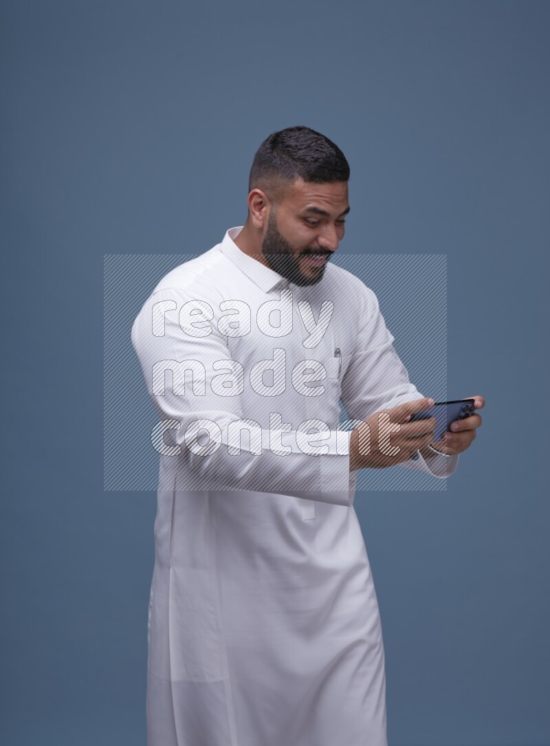 A man Playing a Game on his Smartphone  on Blue Background wearing Saudi Thob