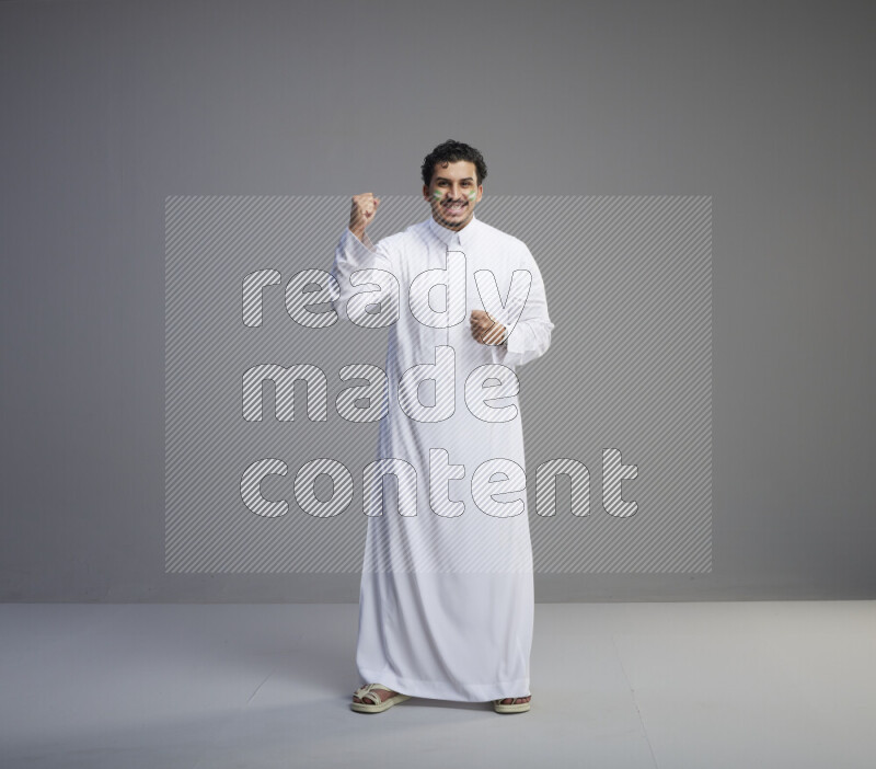 A Saudi man standing wearing thob with face painting interacting with the camera on gray background