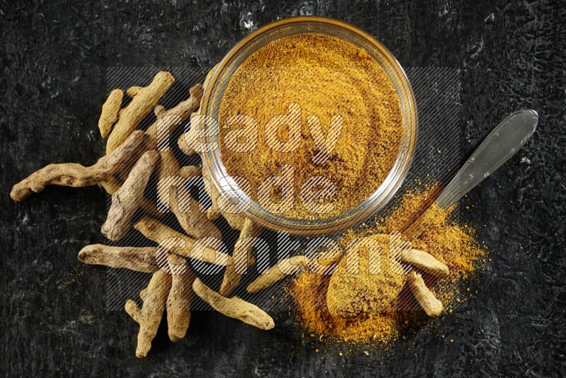 A glass bowl and a metal spoon full of turmeric powder and dried whole fingers next of them on textured black flooring