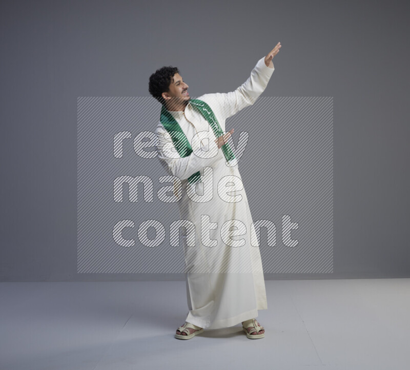 A Saudi man standing wearing thob and Saudi flag scarf on gray background
