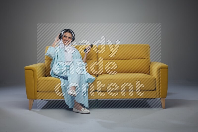 A Saudi woman wearing a light blue Abaya and a white head scarf sitting on a yellow sofa and holding an electronic while wearing headphone eye level on a grey background