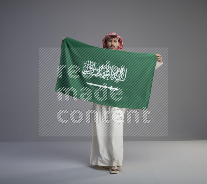 A saudi man standing wearing thob and red shomag holding big saudi flag on gray background
