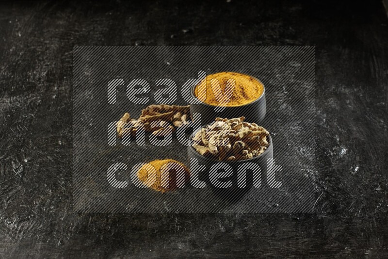 2 pottery black bowls, one full of turmeric powder and the other full of dried turmeric whole fingers on a textured black flooring