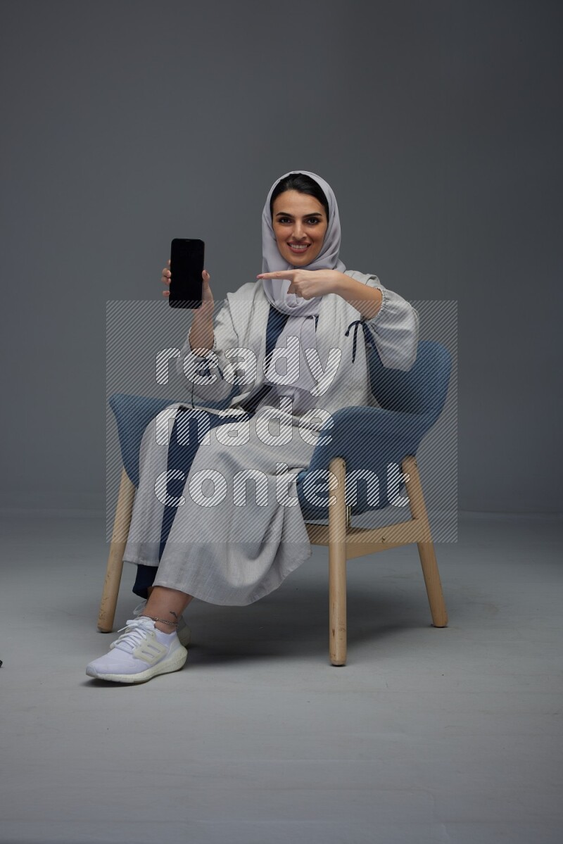 A Saudi woman wearing a light gray Abaya and head scarf sitting on a dark grey chair and showing the phone's screen eye level on a grey background