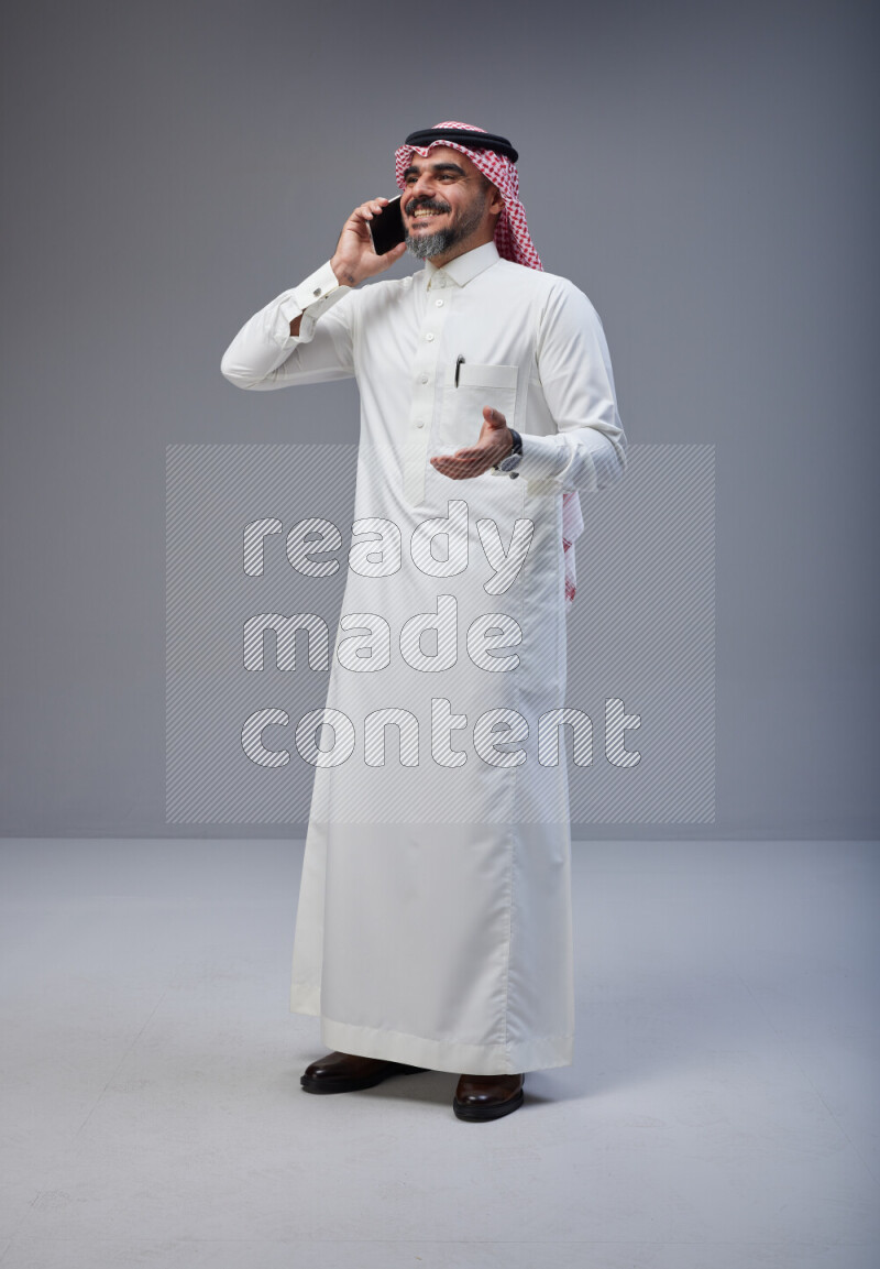 Saudi man Wearing Thob and red Shomag standing talking on phone on Gray background