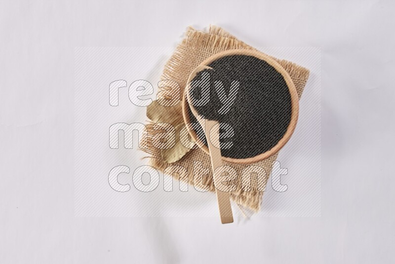 A wooden bowl and wooden spoon full of black seeds on a piece of burlap on a white flooring