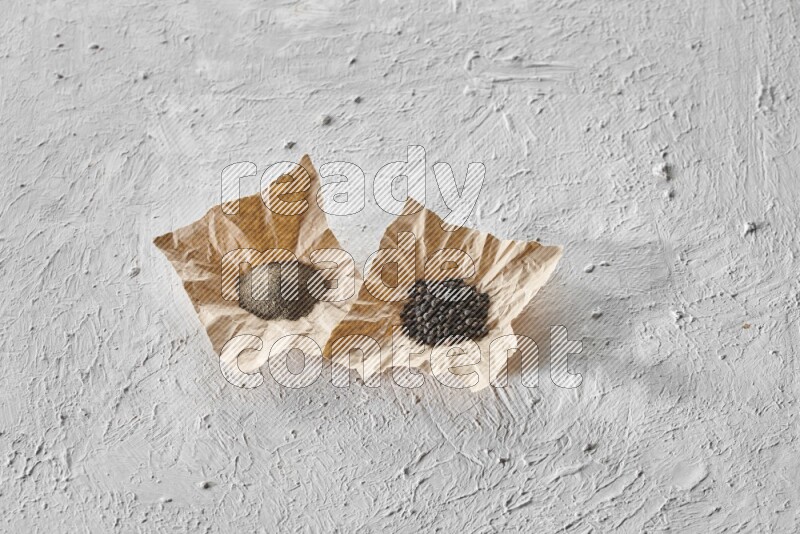 Black pepper and black pepper powder on 2 crumpled pieces of craft paper on a textured white flooring