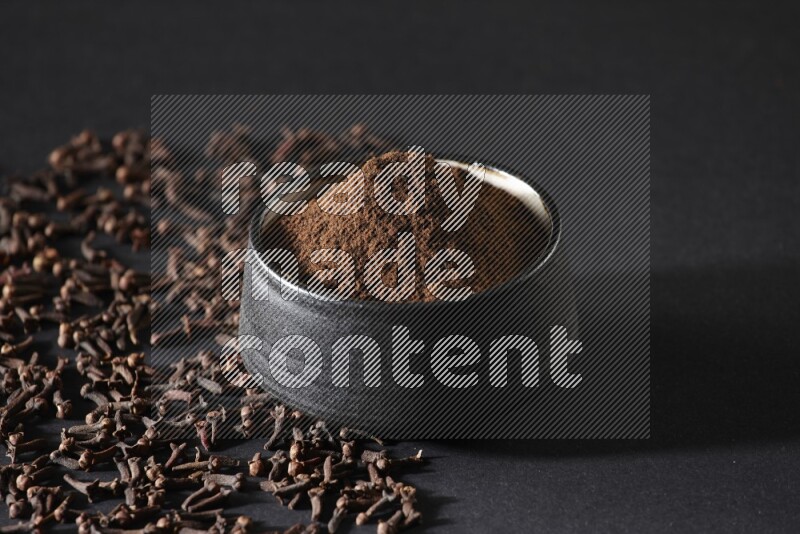 A black pottery bowl full of cloves powder and cloves whole grains on a black flooring