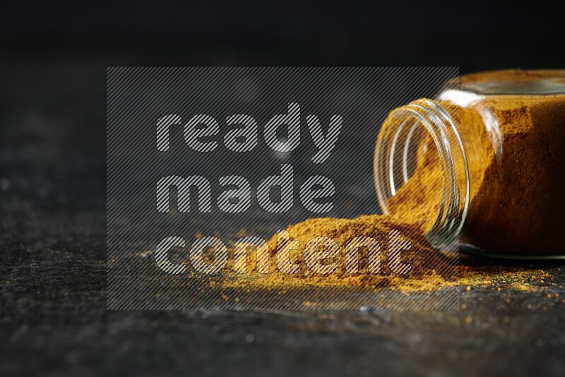A flipped glass spice jar full of turmeric powder and powder spilled out of it on textured black flooring