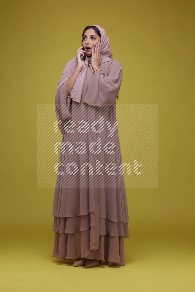 A woman having a call on a Yellow Background wearing Brown Abaya with Hijab