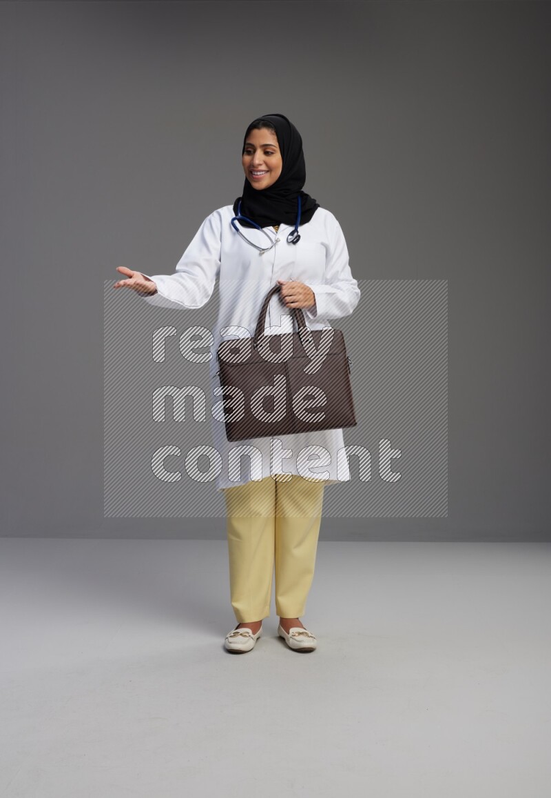 Saudi woman wearing lab coat with stethoscope standing holding bag on Gray background