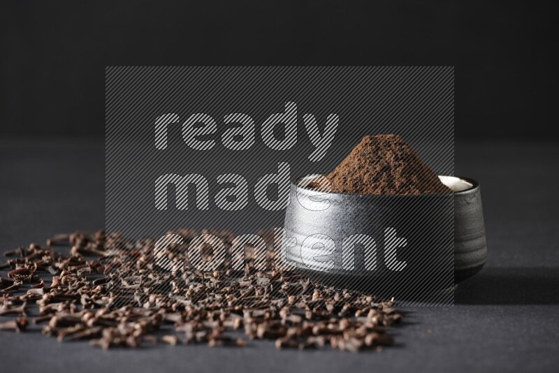 A black pottery bowl full of cloves powder and cloves whole grains on a black flooring