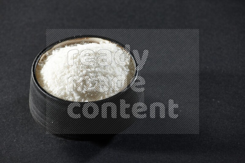 A black pottery bowl full of desiccated coconut on a black background in different angles