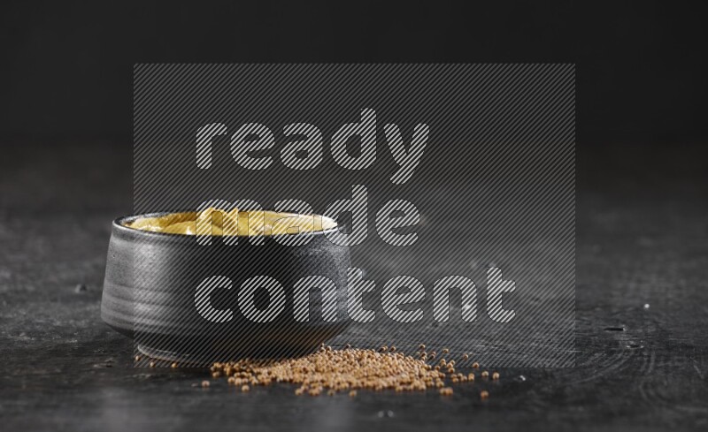 A black pottery bowl full of mustard paste with mustard seeds underneath on textured black flooring