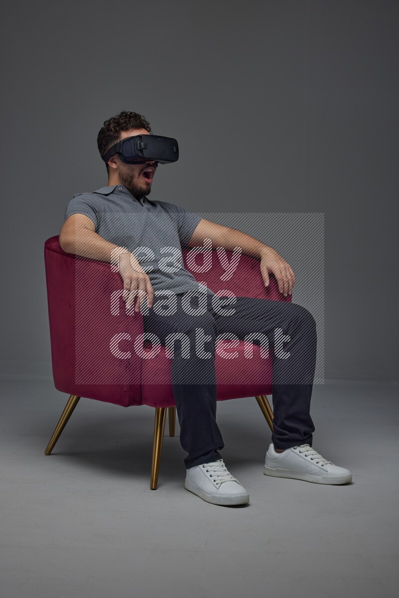 A man wearing casual and wearing VR while sitting on a burgundy chair eye level on a gray background