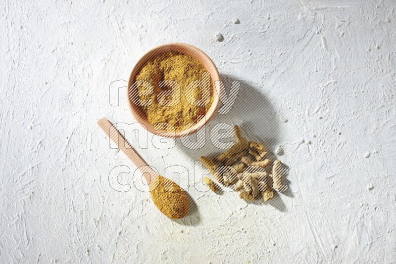 A wooden bowl and wooden spoon full of turmeric powder with dried turmeric fingers on textured white flooring