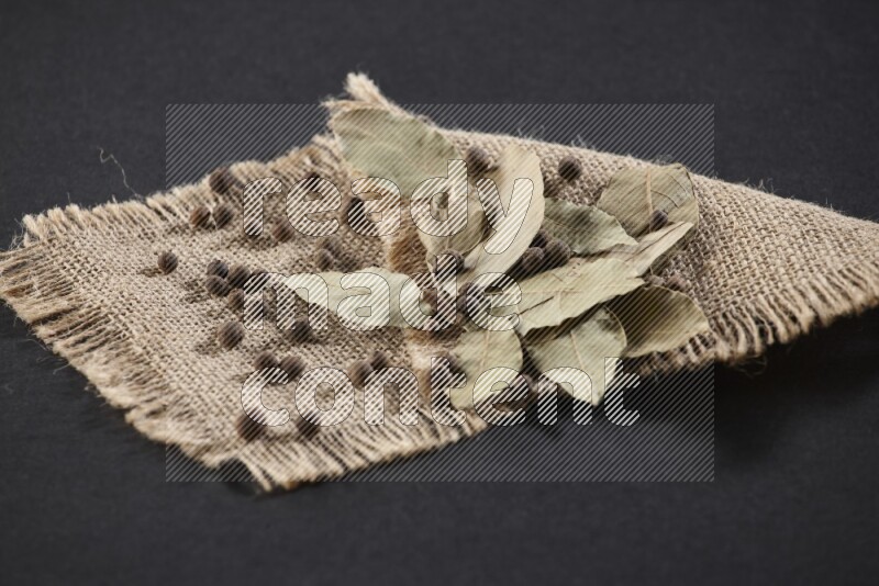 Dried bay leaves and allspice berries on a piece of burlap on black flooring