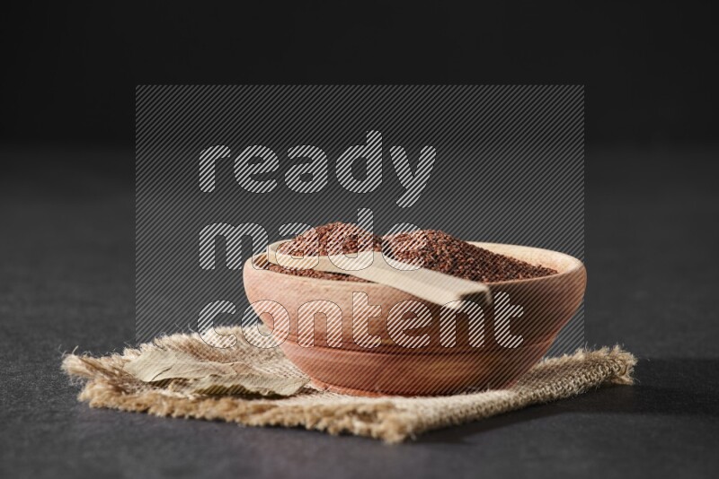 A wooden bowl full of garden cress seeds with wooden spoon full of the seeds on it on burlap fabric on a black flooring