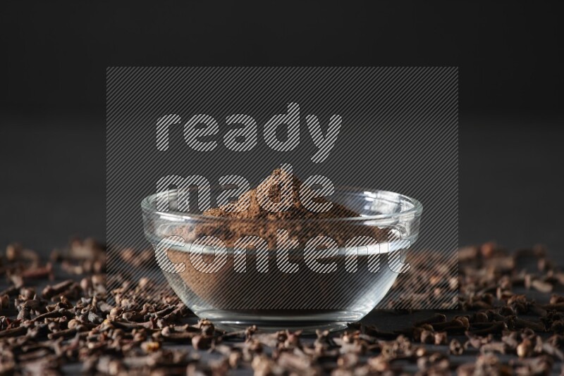 A glass bowl full of cloves powder with cloves grains spread on black flooring