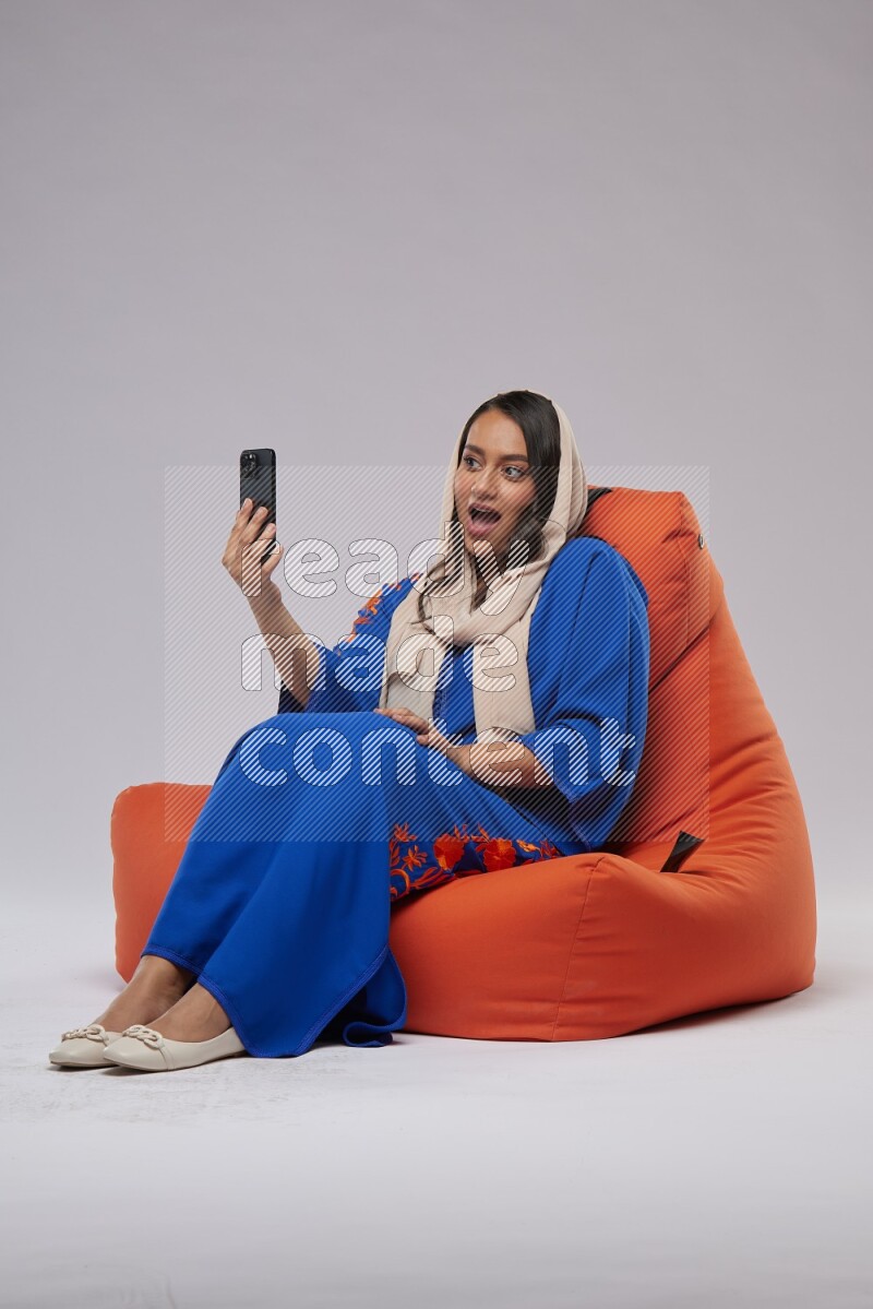A Saudi woman wearing jalabiya sitting on an orange beanbag and taking selfie