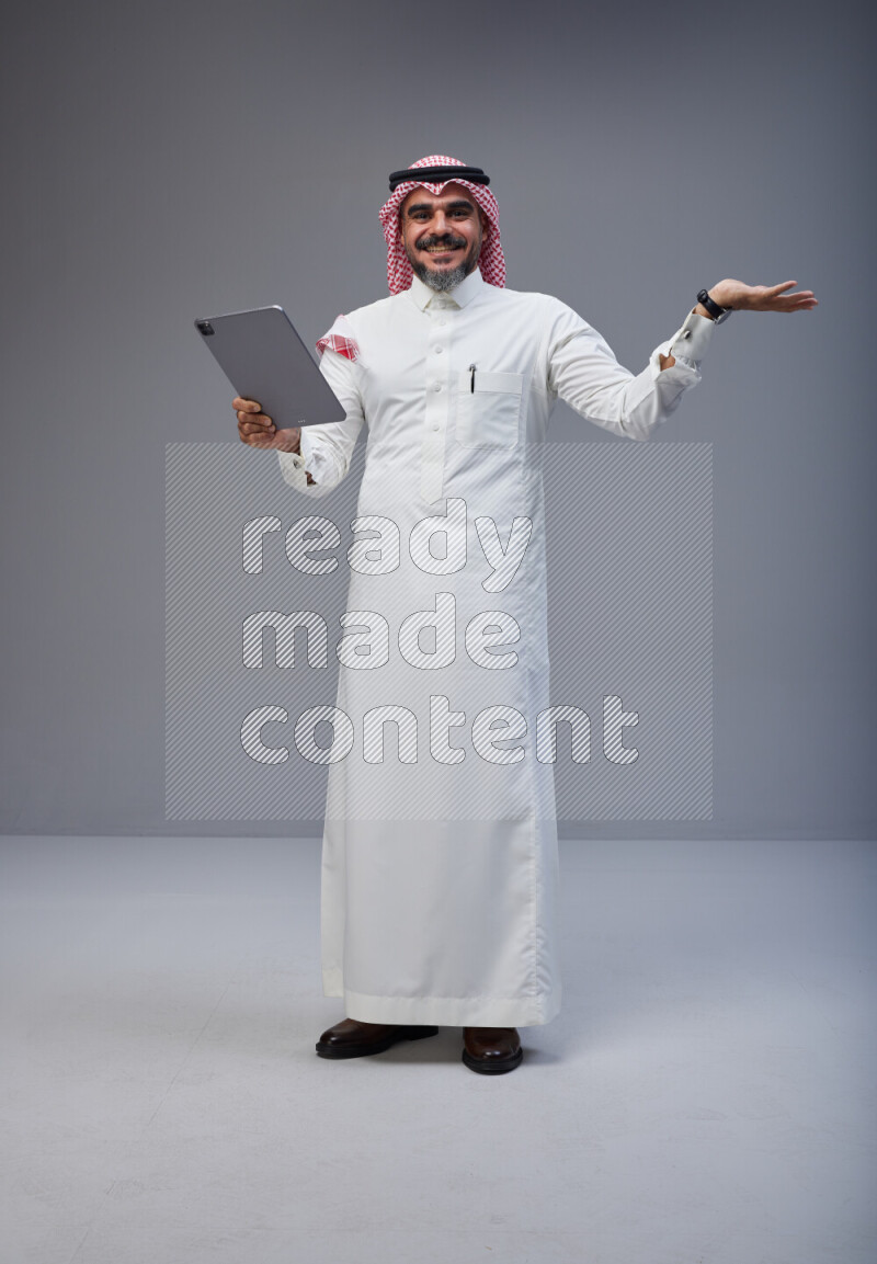 Saudi man Wearing Thob and red Shomag standing working on tablet on Gray background