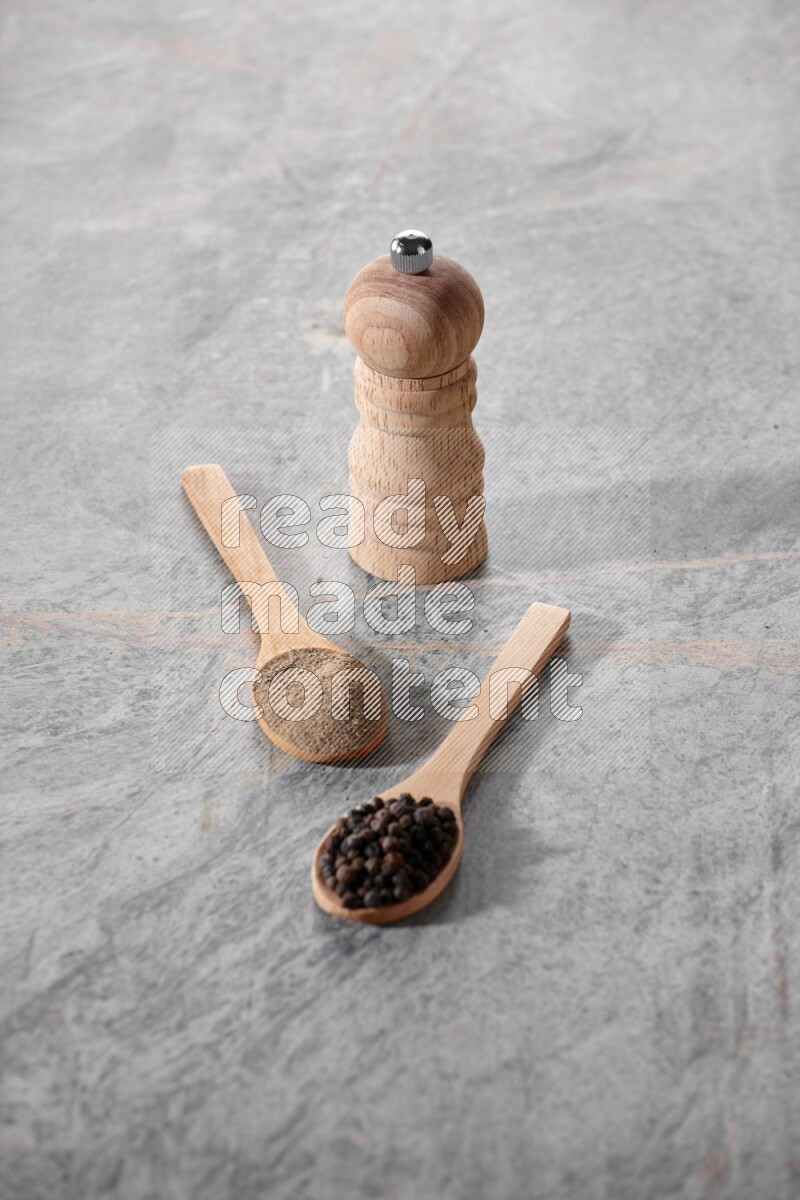 2 wooden spoons, one full of black pepper powder and the other full of black pepper beads and wooden grinder on a marble flooring