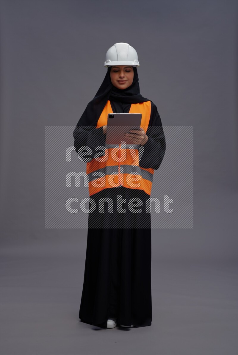 Saudi woman wearing Abaya with engineer vest standing working on tablet on gray background