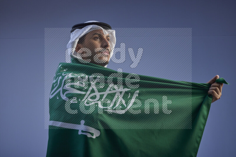 A close-up shot of Saudi man wearing thob and white shomag wrapping big Saudi flag on gray background