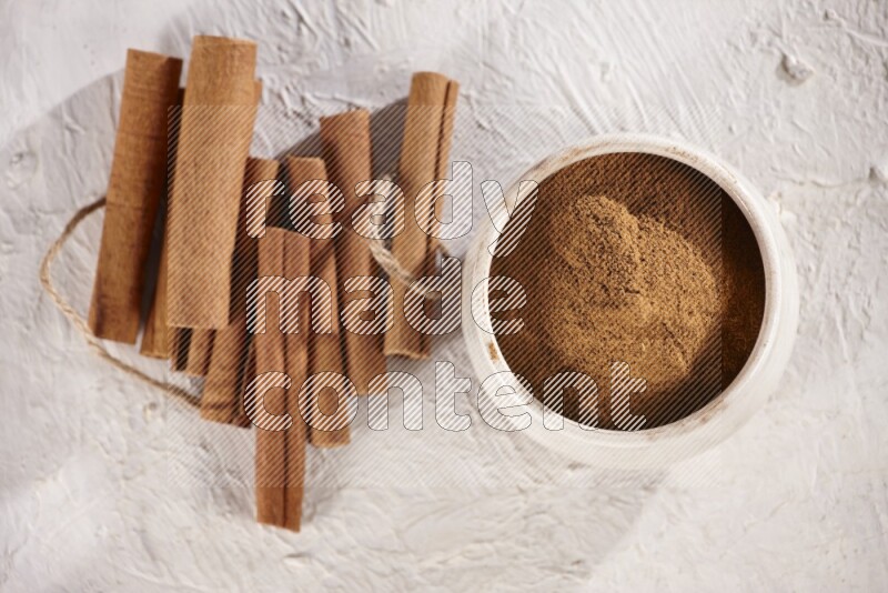 Cinnamon sticks stacked beside a beige bowl full of cinnamon powder on white background