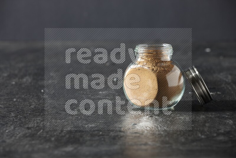 A glass spice jar full of allspice powder on a textured black flooring