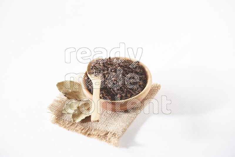 A wooden bowl and a wooden spoon full of cloves on a piece of burlap on a white flooring