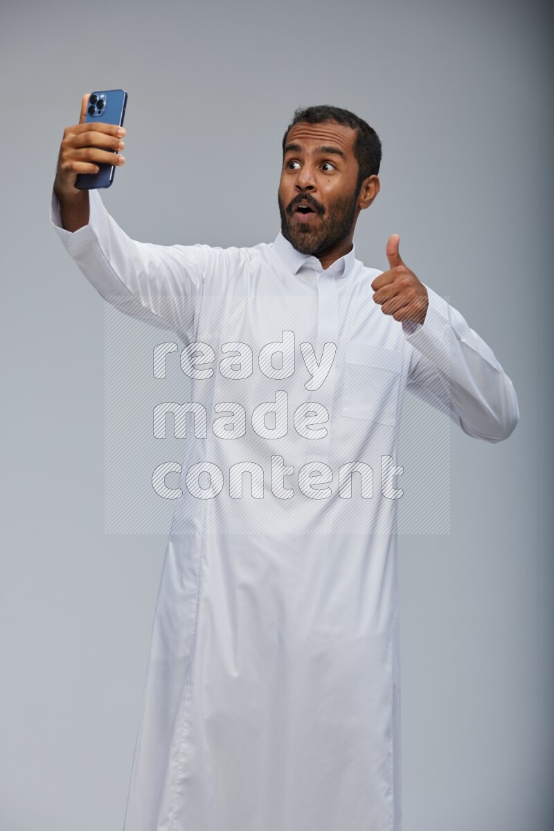 Saudi man wearing Thob standing taking selfie on Gray background