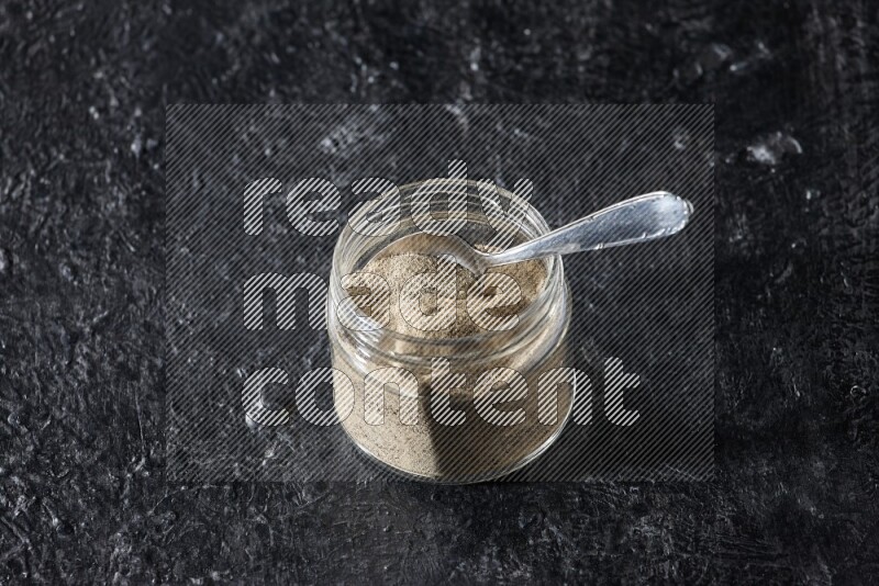 A glass jar and a metal spoon full of white pepper powder on textured black flooring