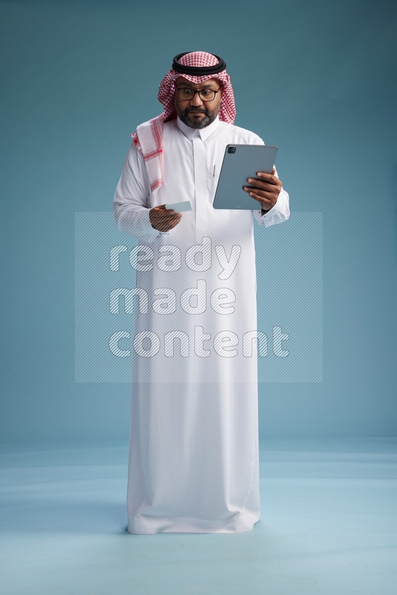 Saudi Man with shimag Standing holding ATM card while working on tablet on blue background