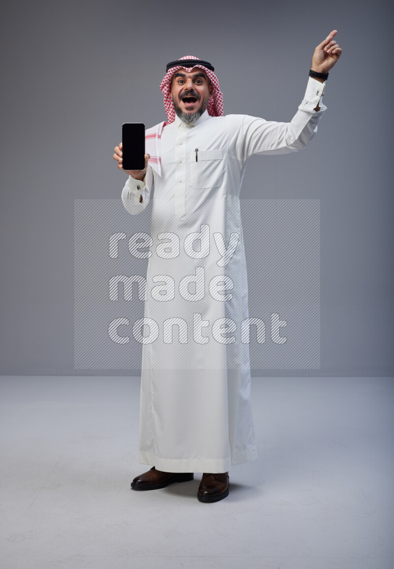 Saudi man Wearing Thob and red Shomag standing showing phone to camera on Gray background