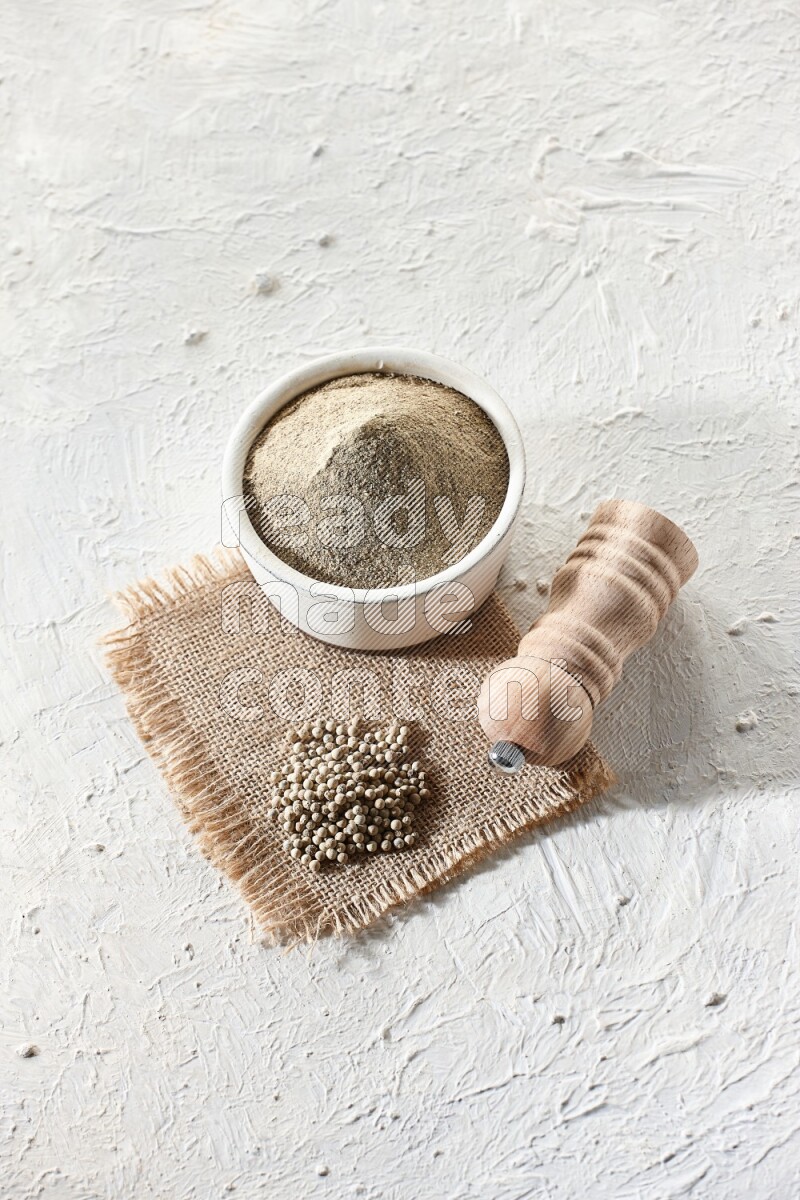 White pottery bowl full of white pepper powder set on a burlap piece of fabric with pepper beads and wooden pepper grinder on textured white flooring