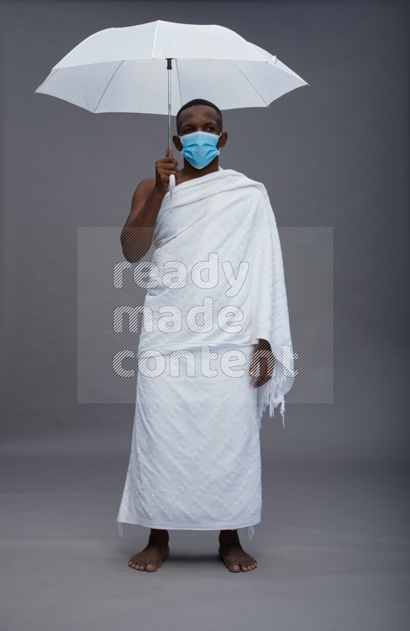 A man wearing Ehram with face mask Standing holding umbrella on gray background