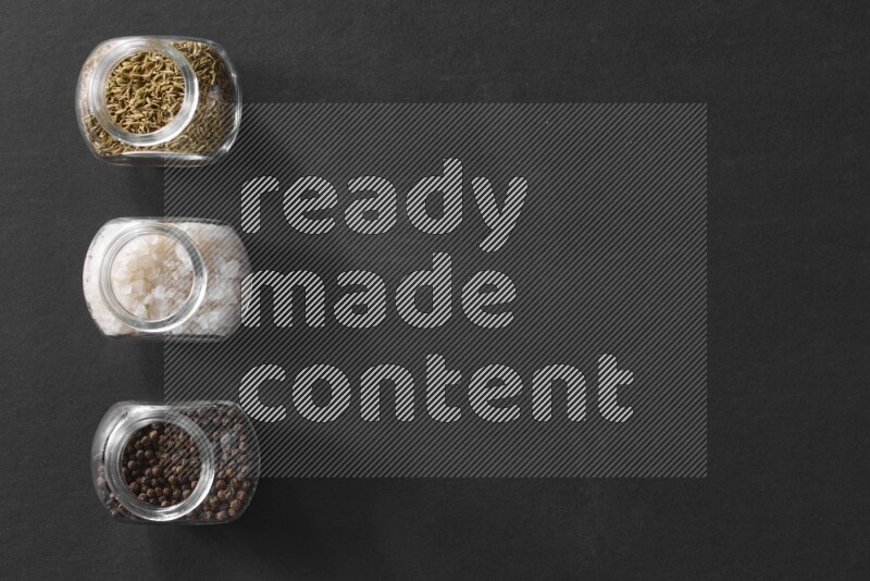 3 glass spice jars full of salt, black peppers and cumin on black flooring