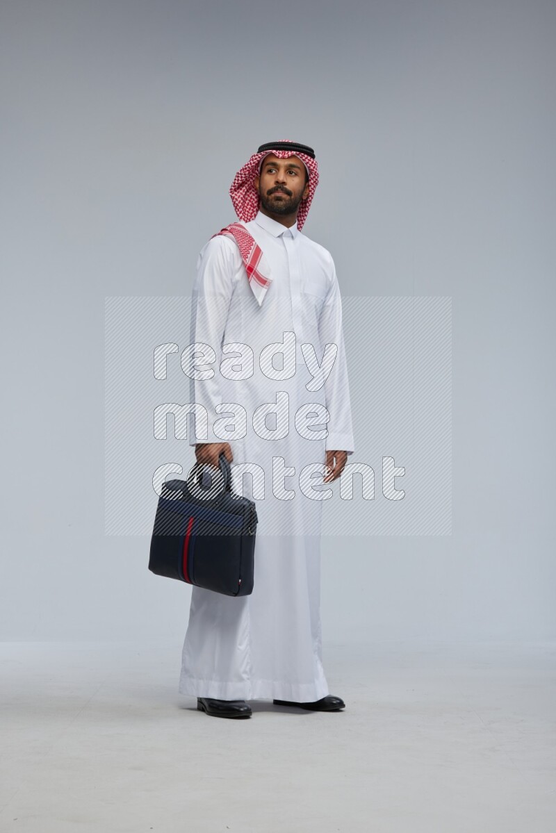 Saudi man Wearing Thob and shomag standing holding bag on Gray background